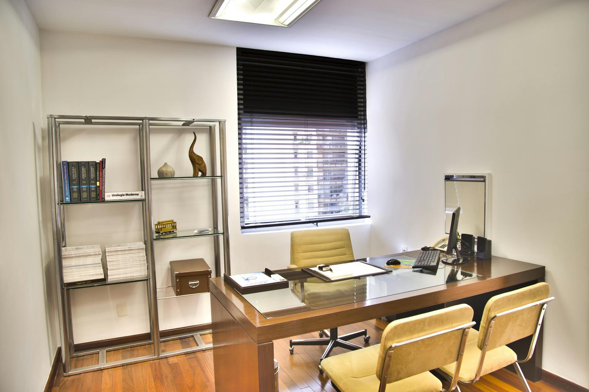 A clean and organised office with yellow chairs, a bookshelf, a window, and stationery and a computer on the desk.